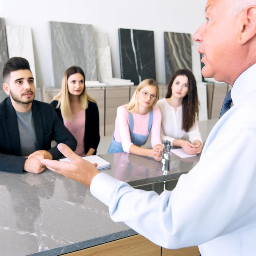 DALL·E 2024-04-22 16.00.49 – A master salesman of marble countertops conducting a training session in a showroom. The experienced salesman, an older gentleman, is demonstrating a